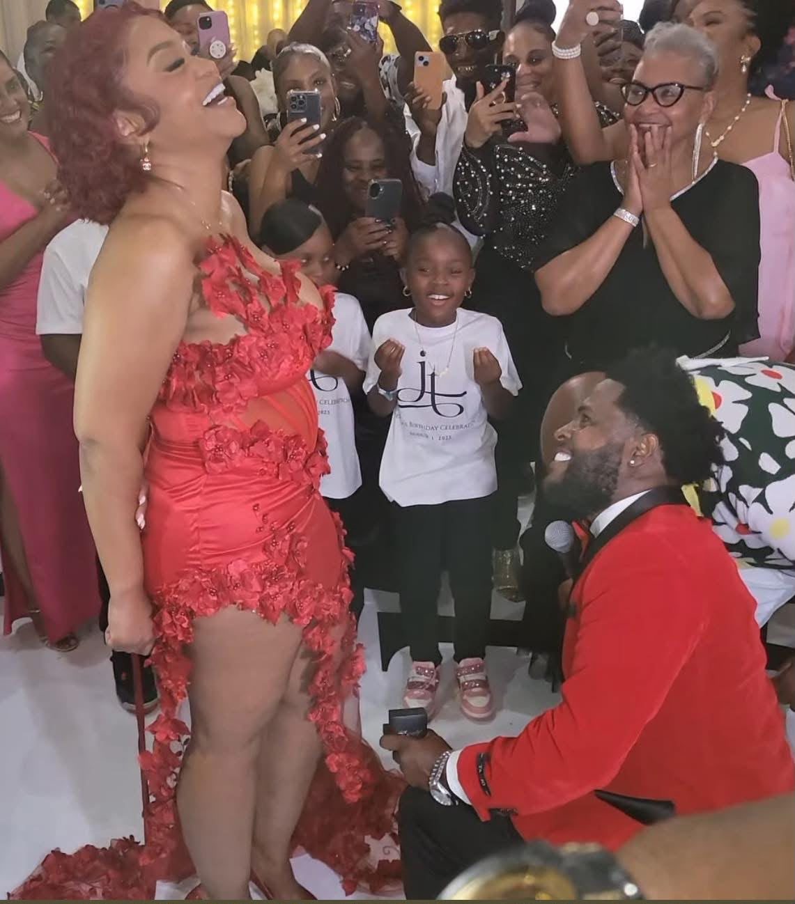 Eugene on one knee holding the ring box as Jessica reacts, surrounded by family and friends